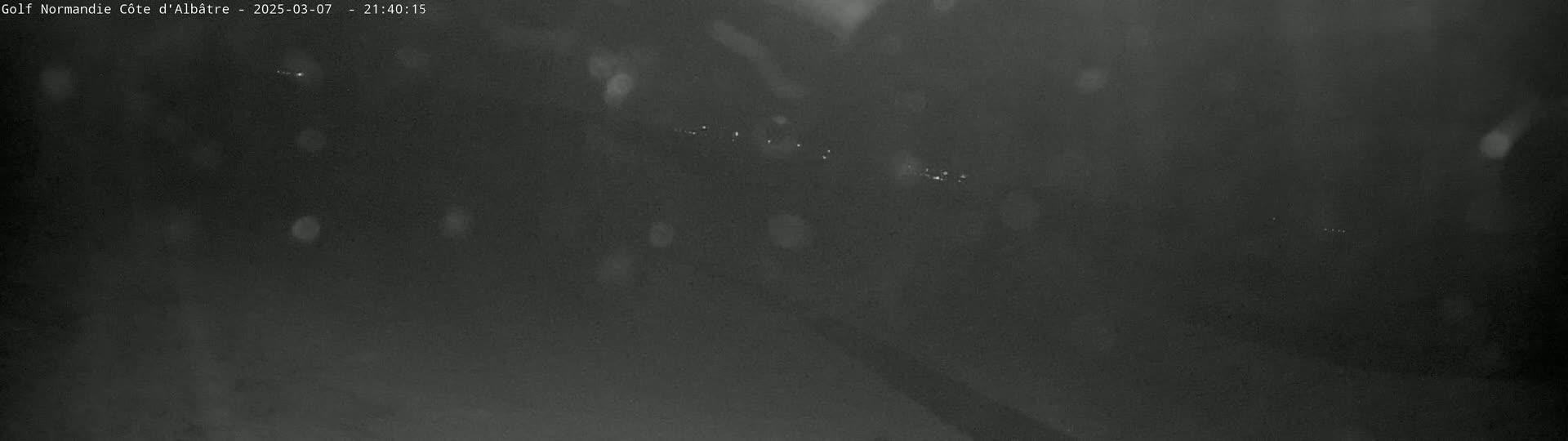 A dark, nighttime scene of a road with faint light sources in the distance, under conditions of light rain or snow.