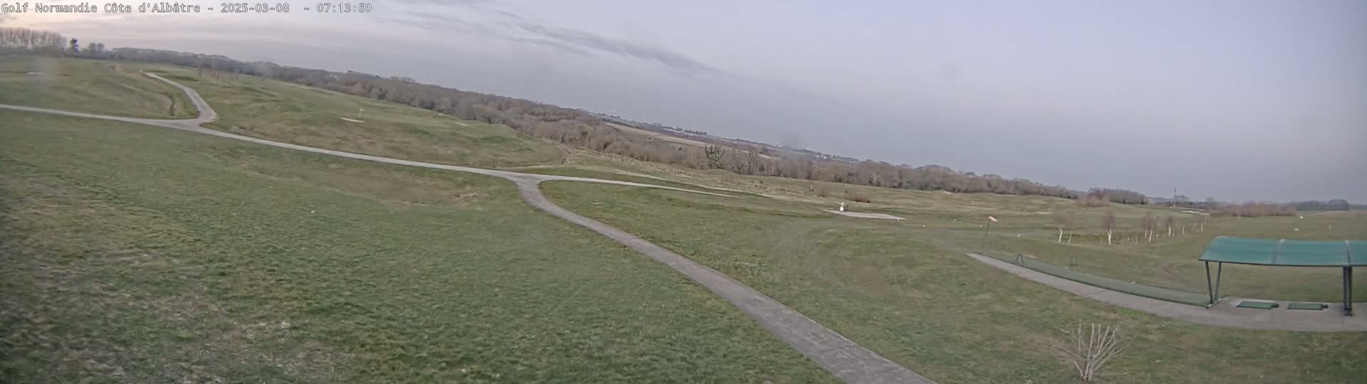 A high-angle view shows a golf course with paths and a covered driving range under an overcast sky.