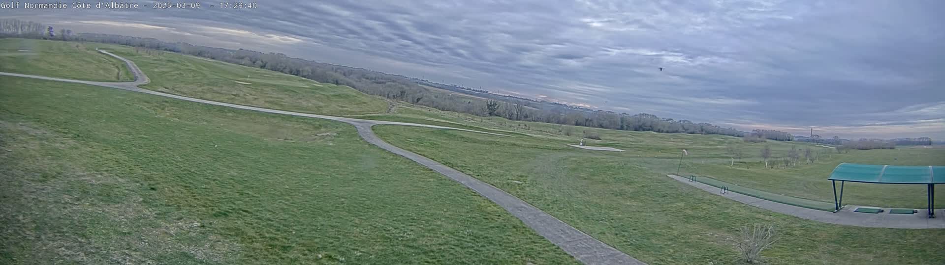 A mostly cloudy sky overlooks a golf course with a paved path and a covered driving range.
