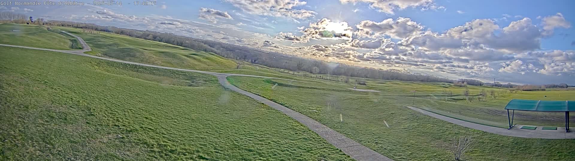 A panoramic view of a golf course on a partly cloudy day, showing rolling green fairways, pathways, and a covered driving range.