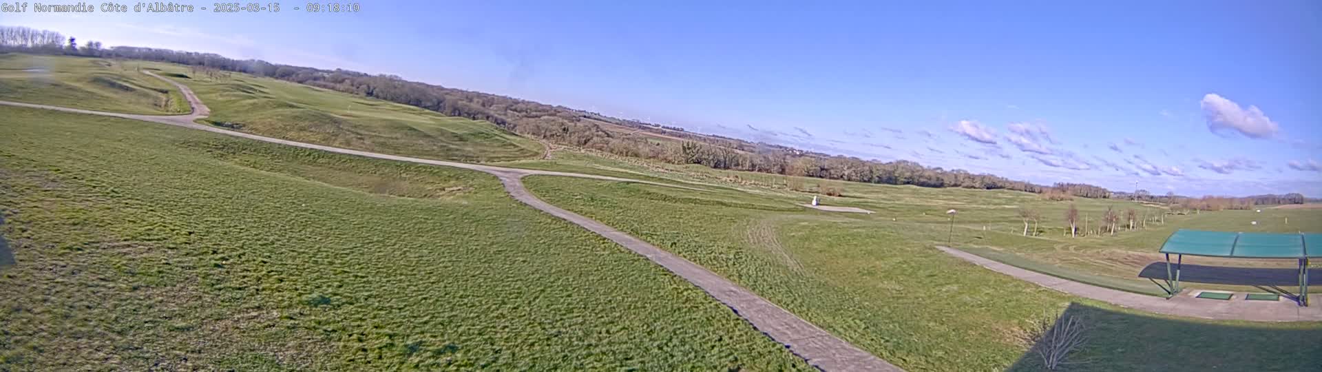 A mostly sunny day reveals a sprawling golf course with a paved path winding through undulating green fairways, and a covered driving range in the foreground.