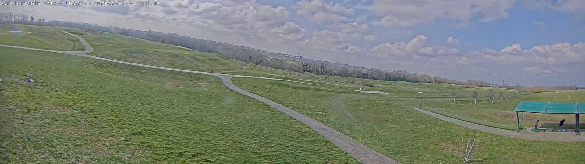 A partly cloudy day at a golf course, showing a wide expanse of grassy fairways, paths, and a driving range with people practicing under a covered area.