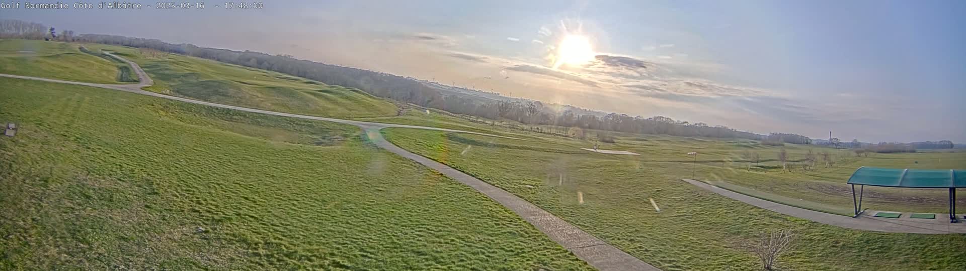 A sunny day on a golf course shows green fairways, winding cart paths, and a covered driving range in the bottom right corner.