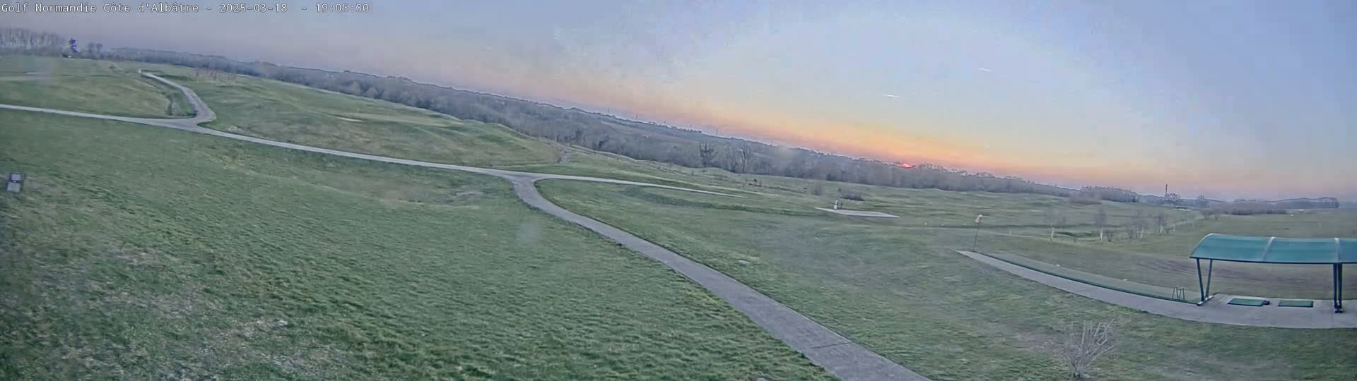 A panoramic view of a golf course at sunset, showing grassy fairways, paved paths, and a covered driving range under a pale, clear sky.