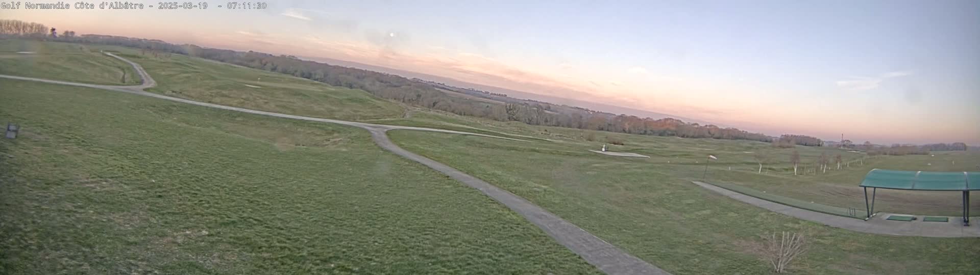 A wide, grassy golf course at dawn under a pale pink and lavender sky, featuring a paved path and a covered driving range.