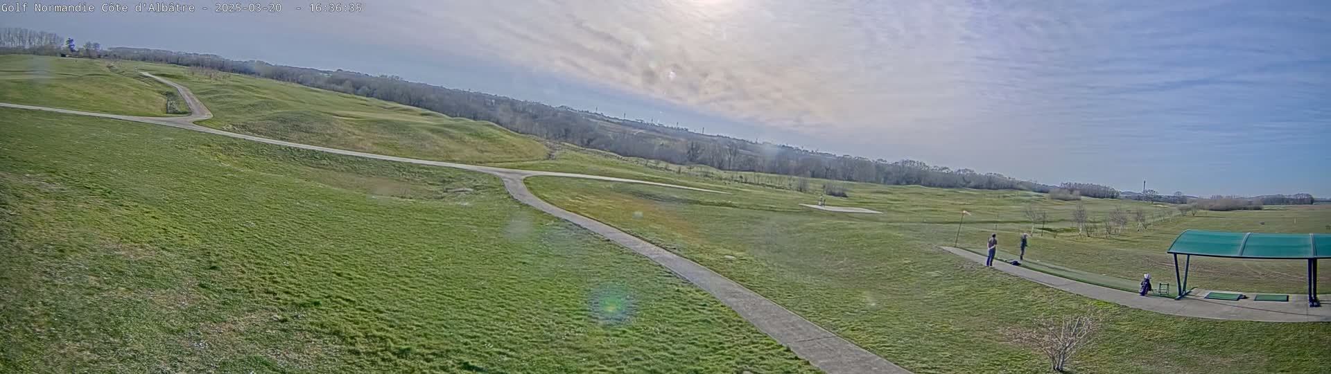 A golf course with a driving range, featuring two people practicing their swings under a partly sunny sky.