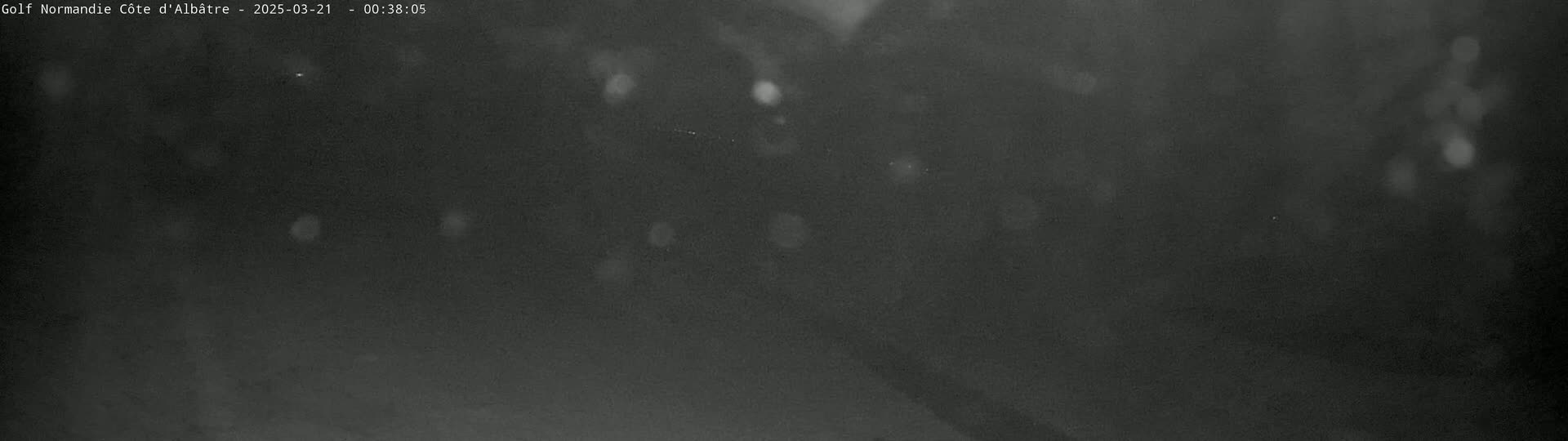 A dark, nighttime outdoor scene shows blurry shapes suggesting trees and possibly a road, with light precipitation visible.