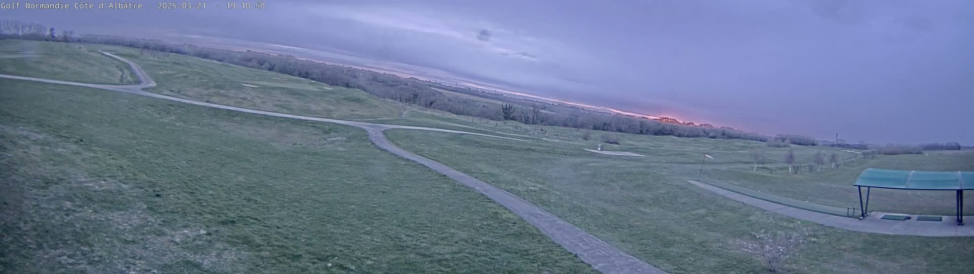 A wide view of a golf course with paved pathways under a twilight sky shows a setting sun on the horizon.