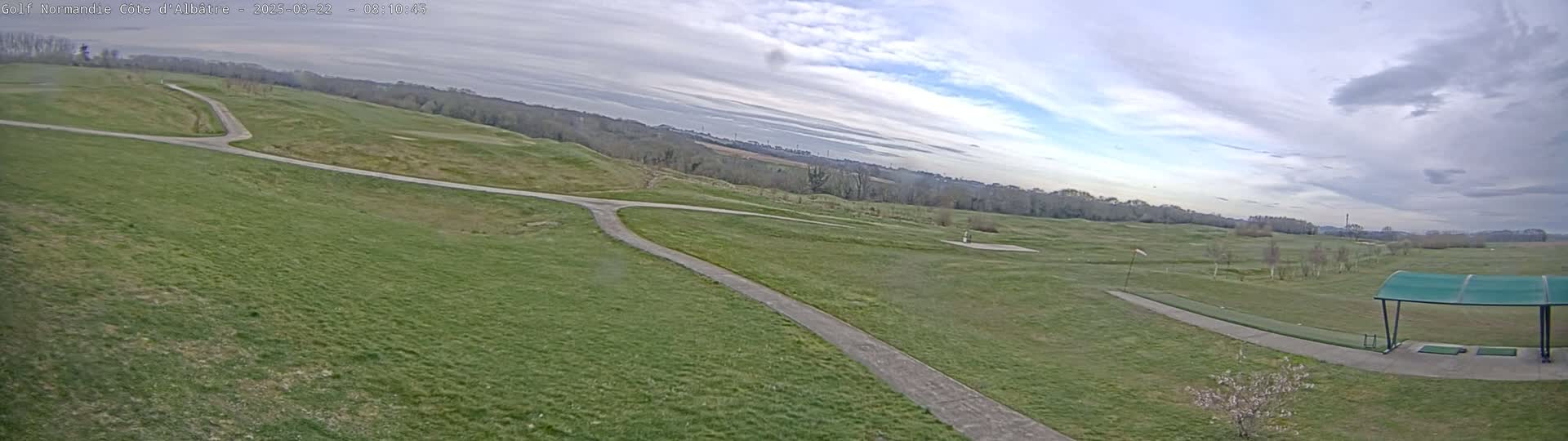 A mostly cloudy day reveals a view of a golf course with paved paths, green fairways, and a covered driving range.