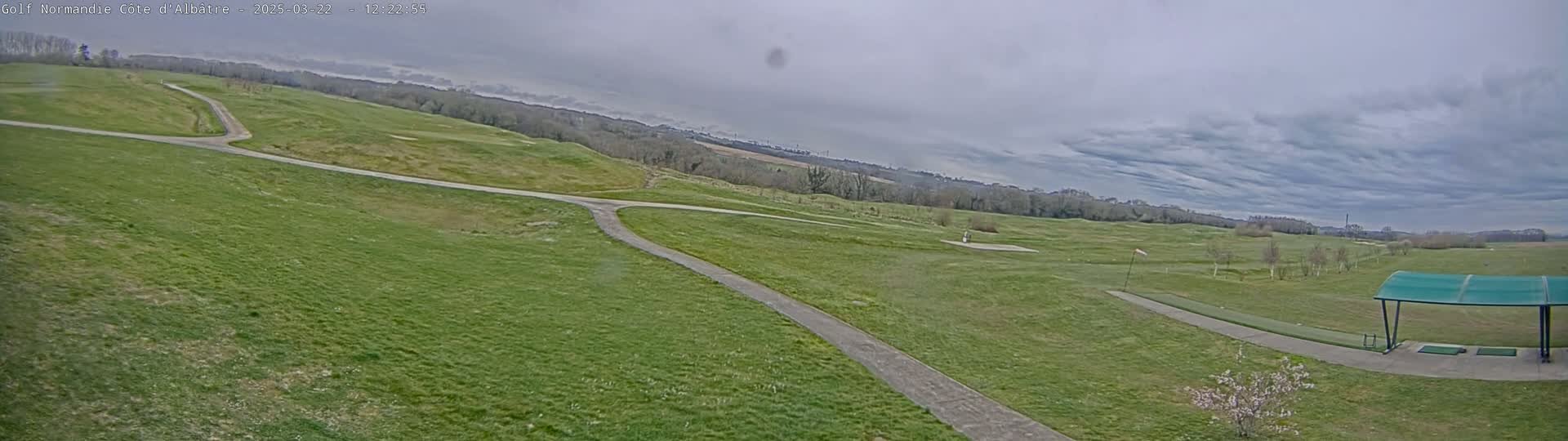 A wide, panoramic view of a golf course with a paved path winding through grassy fairways under a cloudy sky.