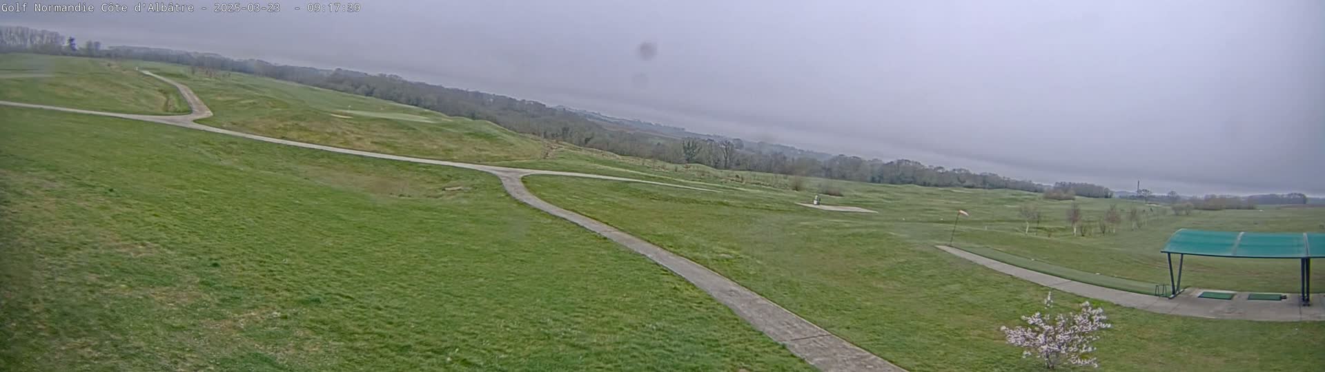 A golf course with paved paths stretches across a gently rolling landscape under an overcast sky.