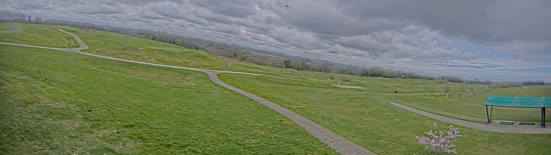 A mostly cloudy day reveals a sprawling green golf course with winding pathways, a driving range with a covered area, and a distant horizon of trees under a partly cloudy sky.