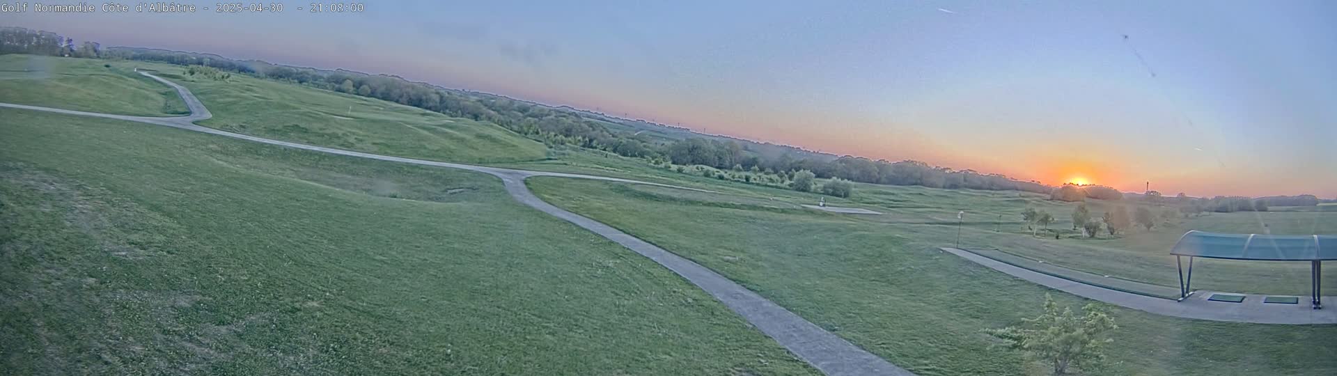 A panoramic view of a golf course at sunset, with a paved path winding through the green grass and a covered driving range in the foreground.