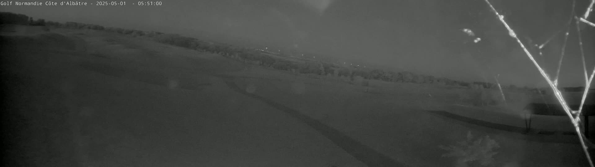 A nighttime, low-light image shows a mostly flat, snow-covered landscape with sparse trees and shrubs in the background under a hazy sky.
