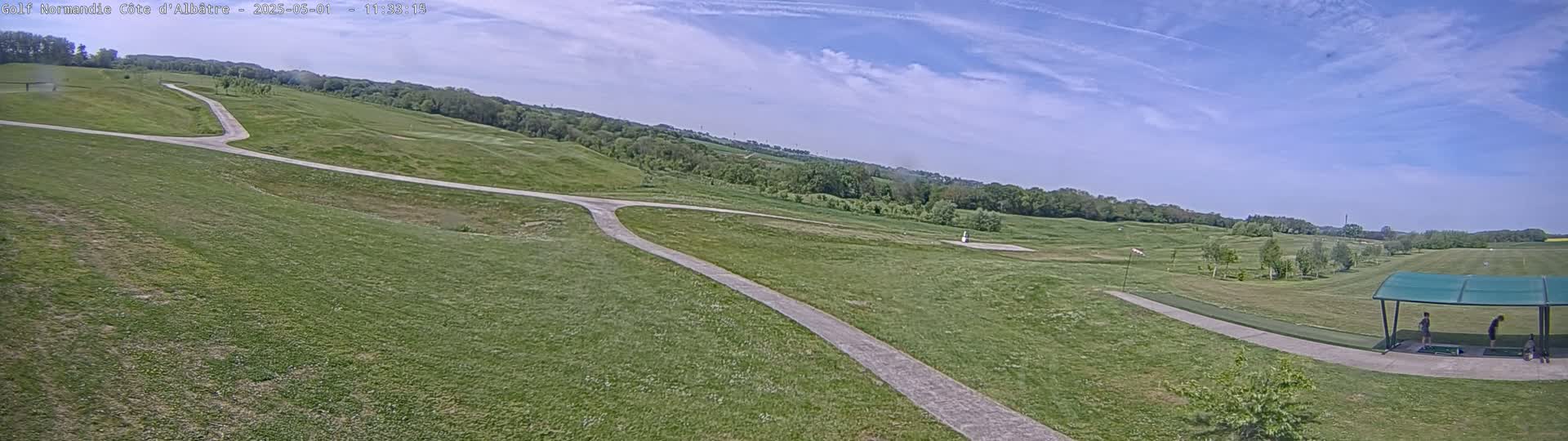 A mostly sunny day overlooks a golf course with paved paths winding through grassy hills, and a covered driving range with a few people.
