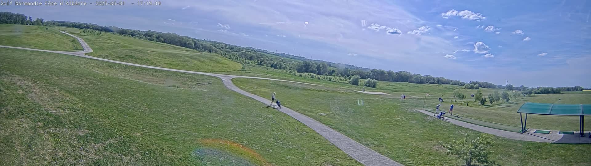 A golf course with a few people walking and golfing under a partly sunny sky.
