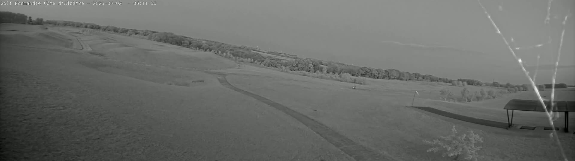 A grayscale image shows a golf course with rolling hills, sparse trees, and a covered area in the lower right corner under an overcast sky.