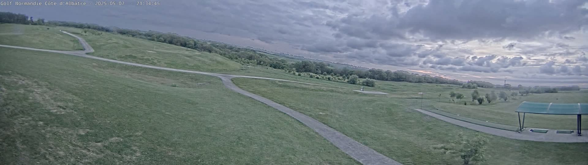 A golf course with paved pathways winds through grassy hills under a cloudy sky.