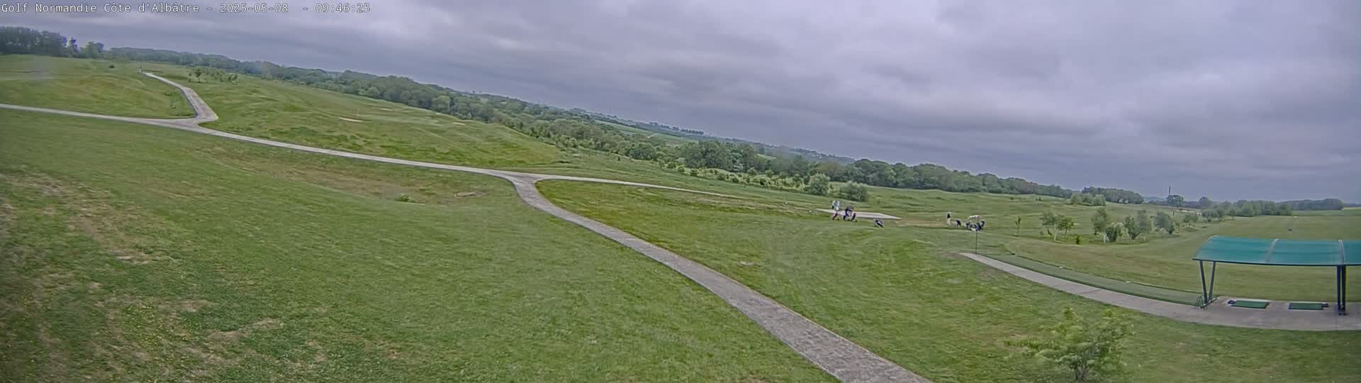 A golf course with a paved path, under a cloudy sky, shows several golfers on the course near a covered driving range.