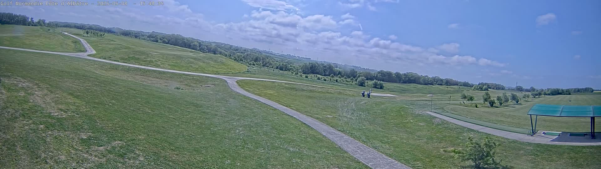 A mostly sunny day shows a view of a golf course with a paved path winding through the green fairways, a covered driving range, and a few people in the distance.