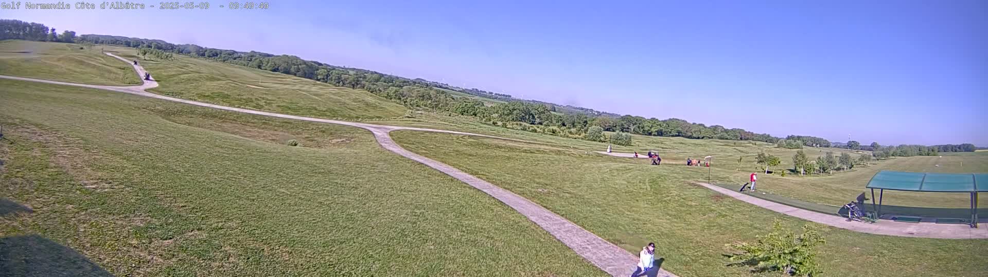 A sprawling golf course with paved pathways winds through rolling green hills under a clear blue sky.