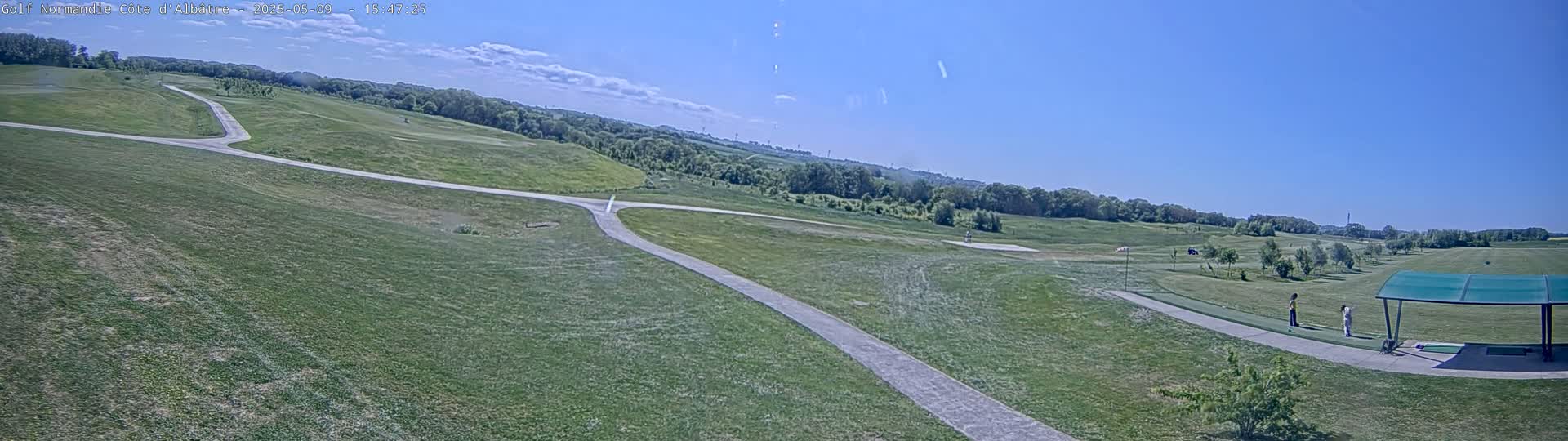 A mostly sunny day reveals a sprawling golf course with paved paths, a driving range with golfers under a covered area, and a distant tree line under a clear blue sky.