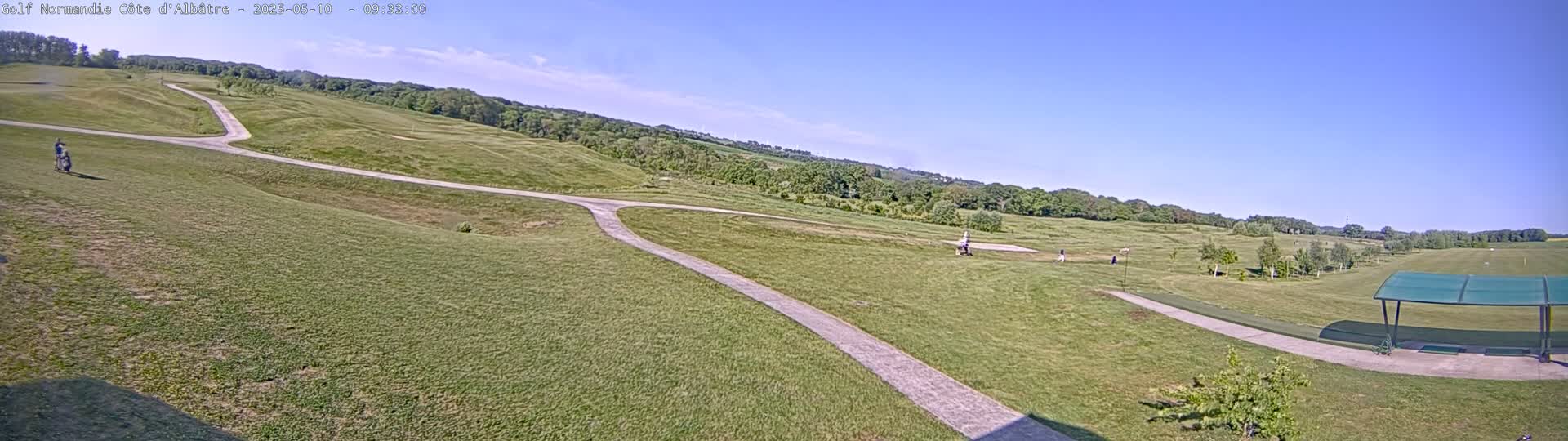 A mostly sunny day at a golf course shows a wide expanse of green grass, a paved path winding through the fairways, and a few golfers visible in the distance near a covered driving range.