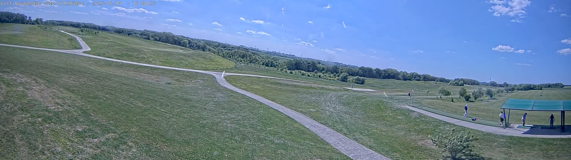 A panoramic view of a golf course on a partly sunny day shows a paved path winding through grassy fairways, with several golfers visible near a covered driving range.