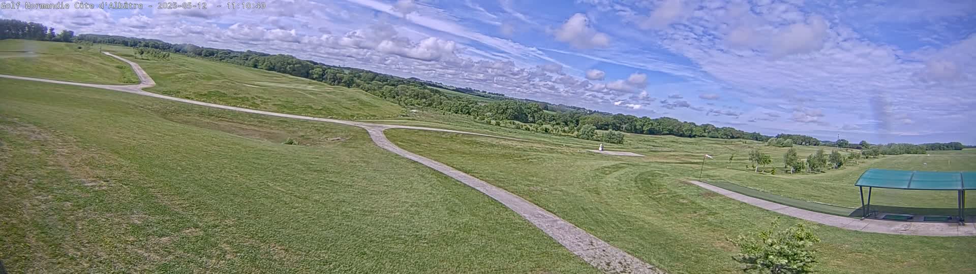 A wide, high-angle view shows a grassy golf course with paved paths under a partly cloudy sky.