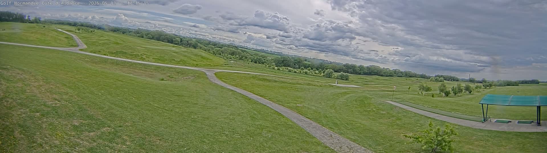 A panoramic view of a golf course on a partly cloudy day shows a paved path winding through a grassy area with a driving range and covered hitting bays in the foreground.