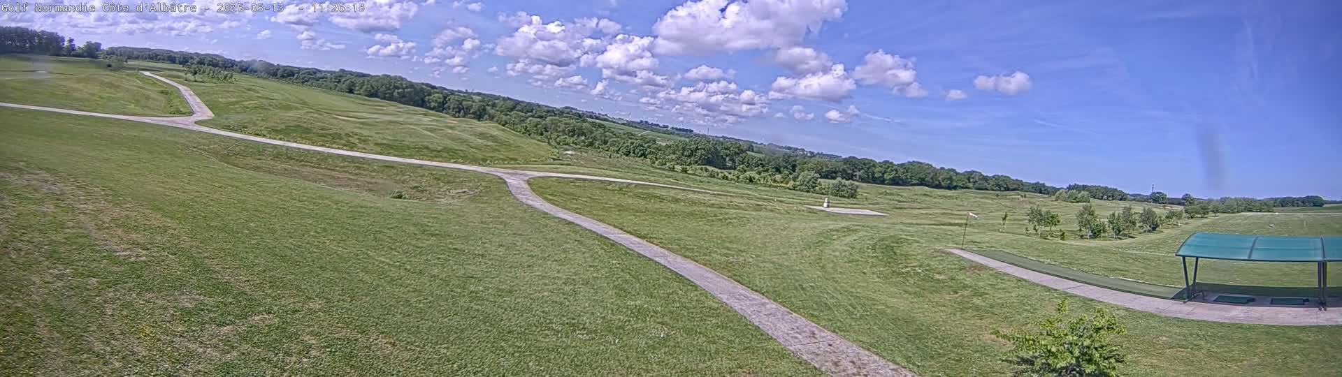 A grassy golf course with a paved path, under a partly cloudy blue sky.