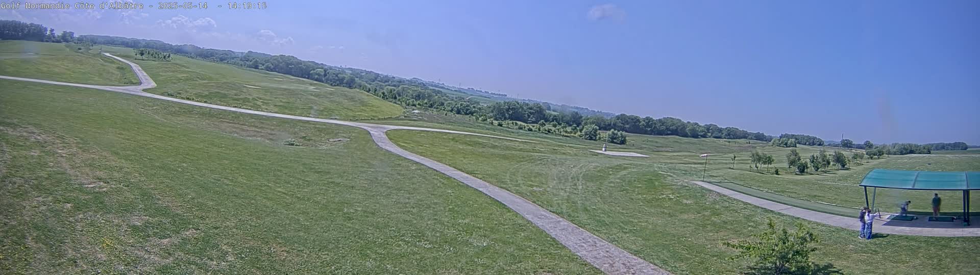 A panoramic view of a golf course on a sunny day shows a paved path splitting into two directions across a grassy expanse with trees in the background.
