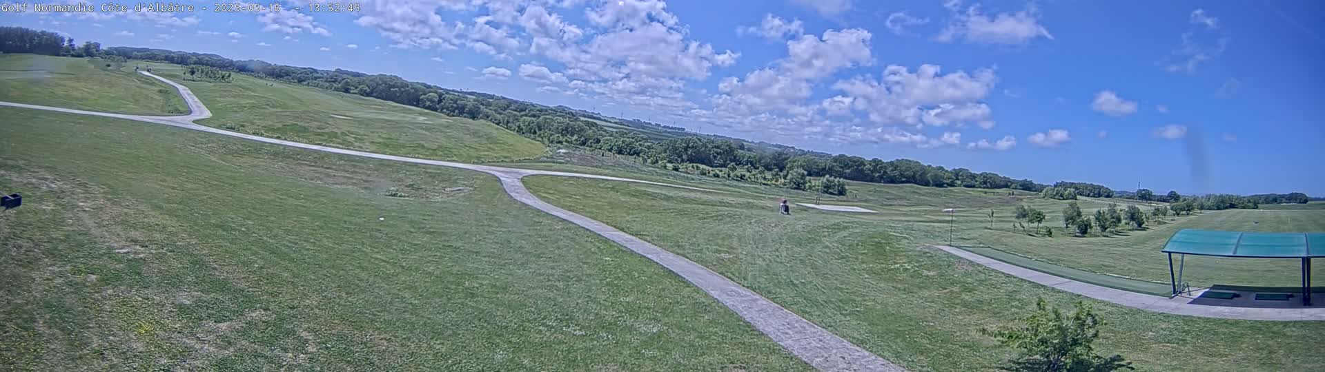 A panoramic view of a golf course on a partly cloudy day, showing a paved path winding through grassy fairways and greens, with a covered driving range in the foreground.
