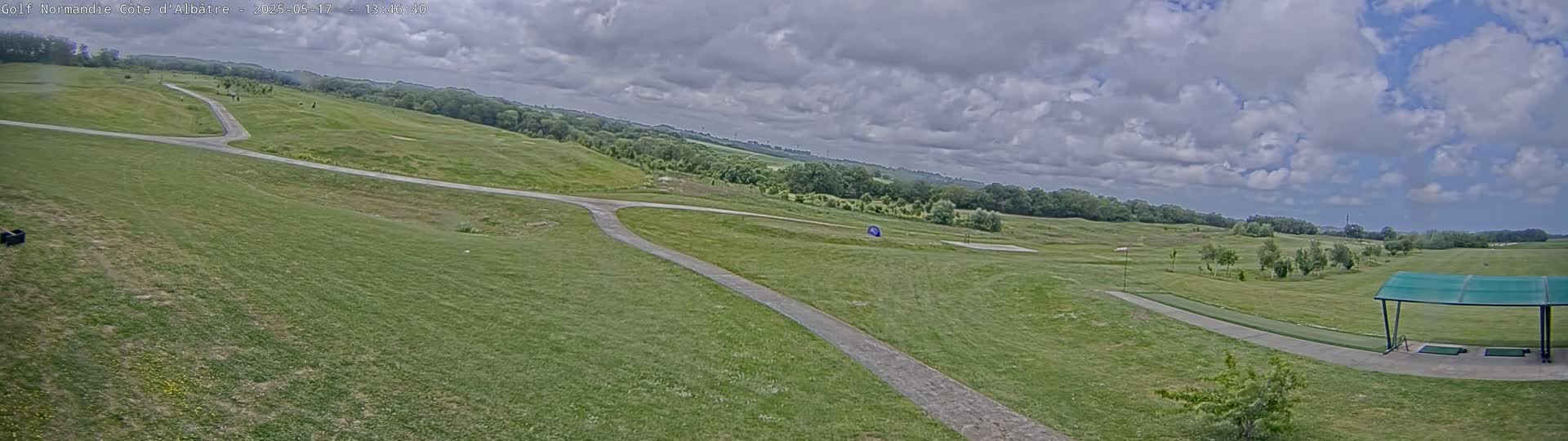 A partly cloudy day at a golf course shows a grassy landscape with rolling hills, paved paths, and a covered driving range.