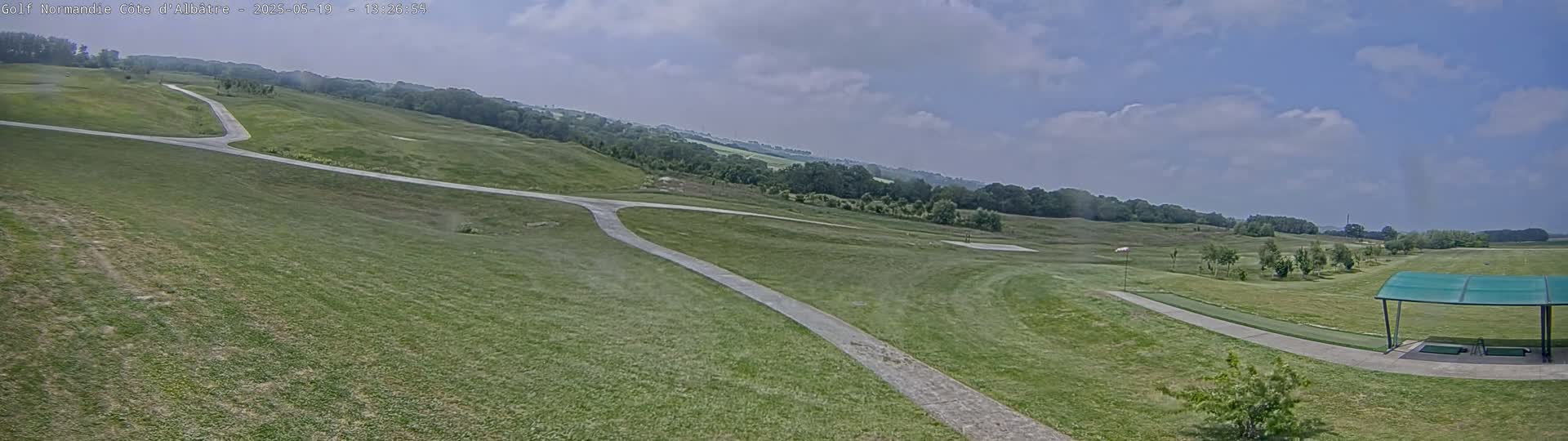 A panoramic view of a golf course on a partly cloudy day shows a paved path winding through grassy fairways and a covered driving range in the foreground.