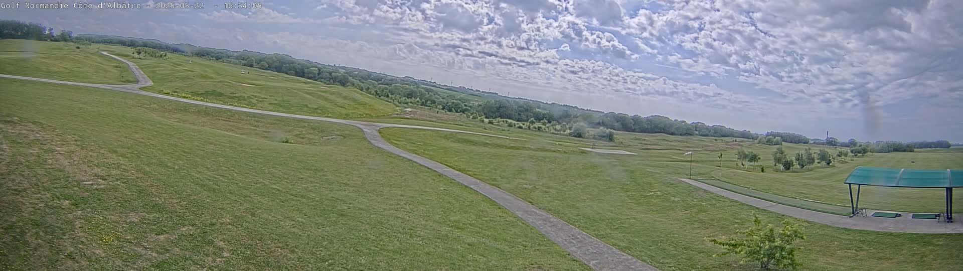 A partly cloudy sky overlooks a golf course with a paved path and a covered driving range.