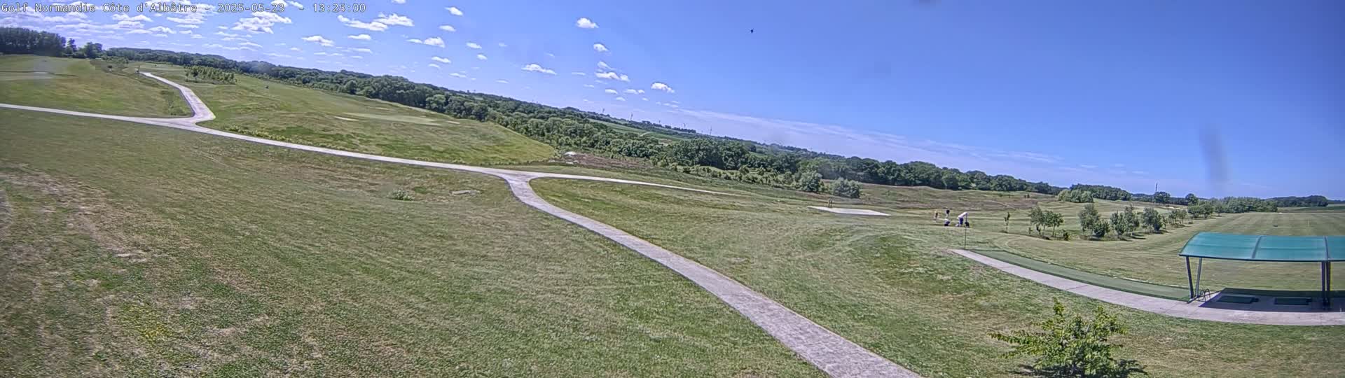 A sunny day on a golf course shows a paved path winding through grassy fairways, with golfers visible in the distance and a covered shelter near the bottom of the frame.