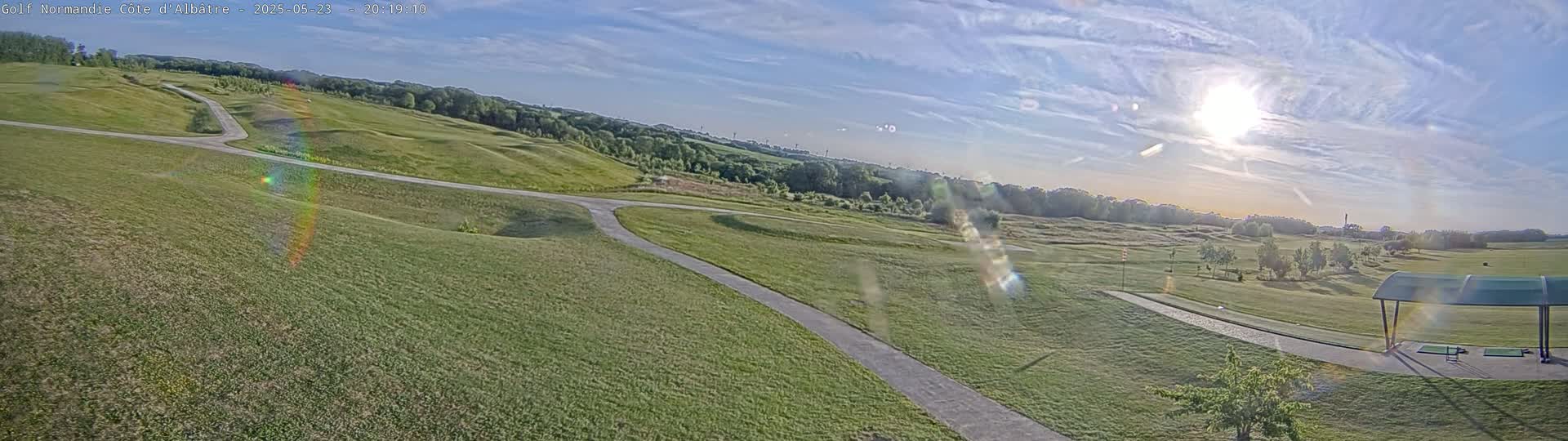 A sunny day shows a golf course with rolling green hills, paths, and a covered driving range.