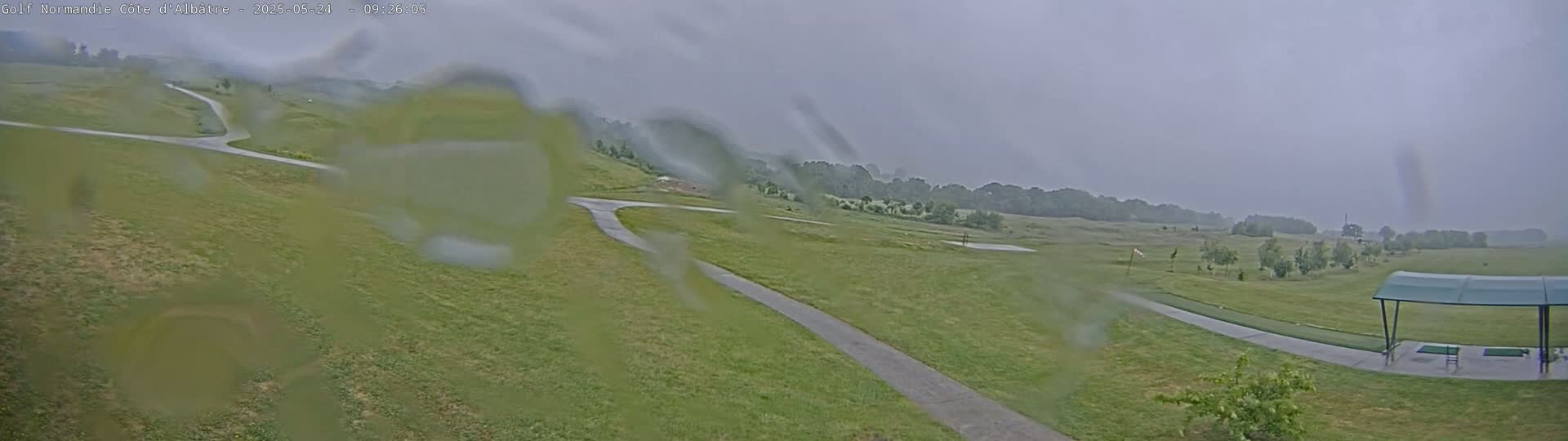 A rain-covered view of a golf course with winding cart paths and a covered driving range in the background.