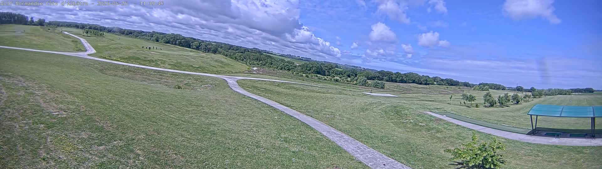 A partly sunny day reveals a sprawling golf course with winding paved paths, a driving range with covered stalls, and a background of verdant trees under a partly cloudy sky.