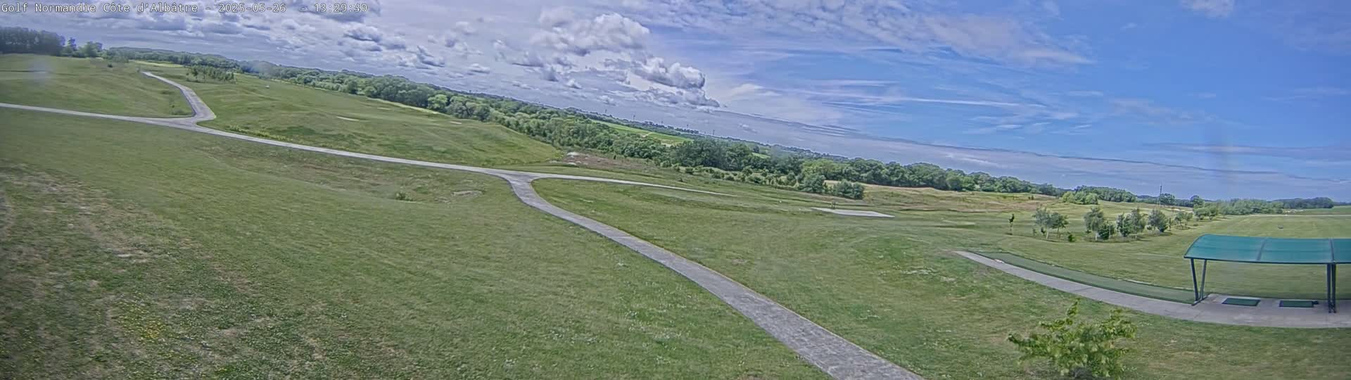 A panoramic view of a golf course on a partly cloudy day shows a paved path winding through a grassy area, leading to a driving range with a covered shelter.