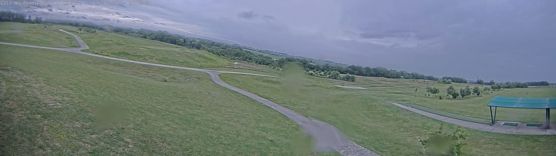 A wide, panoramic view of a golf course under a cloudy sky shows a paved path winding through grassy fairways and a driving range with a covered area.