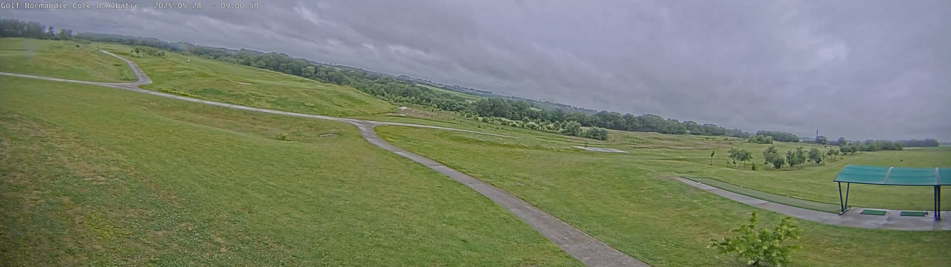 A mostly overcast sky hangs over a sprawling, green golf course with a paved path winding through it, leading to a covered driving range.