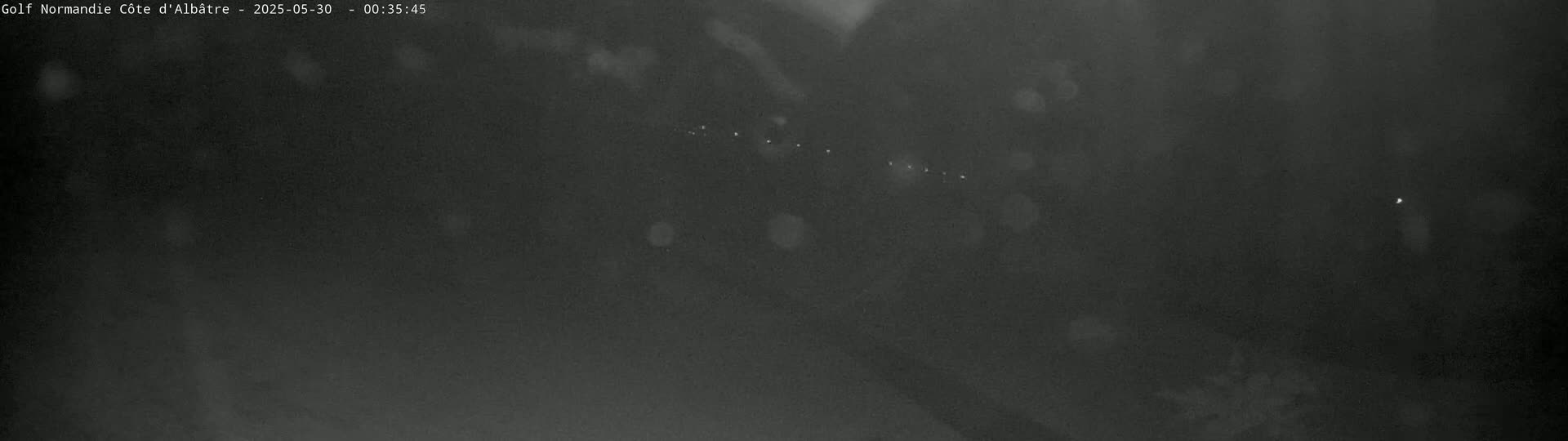 A dark, nighttime scene shows a distant line of faint lights, possibly headlights, through a hazy atmosphere.
