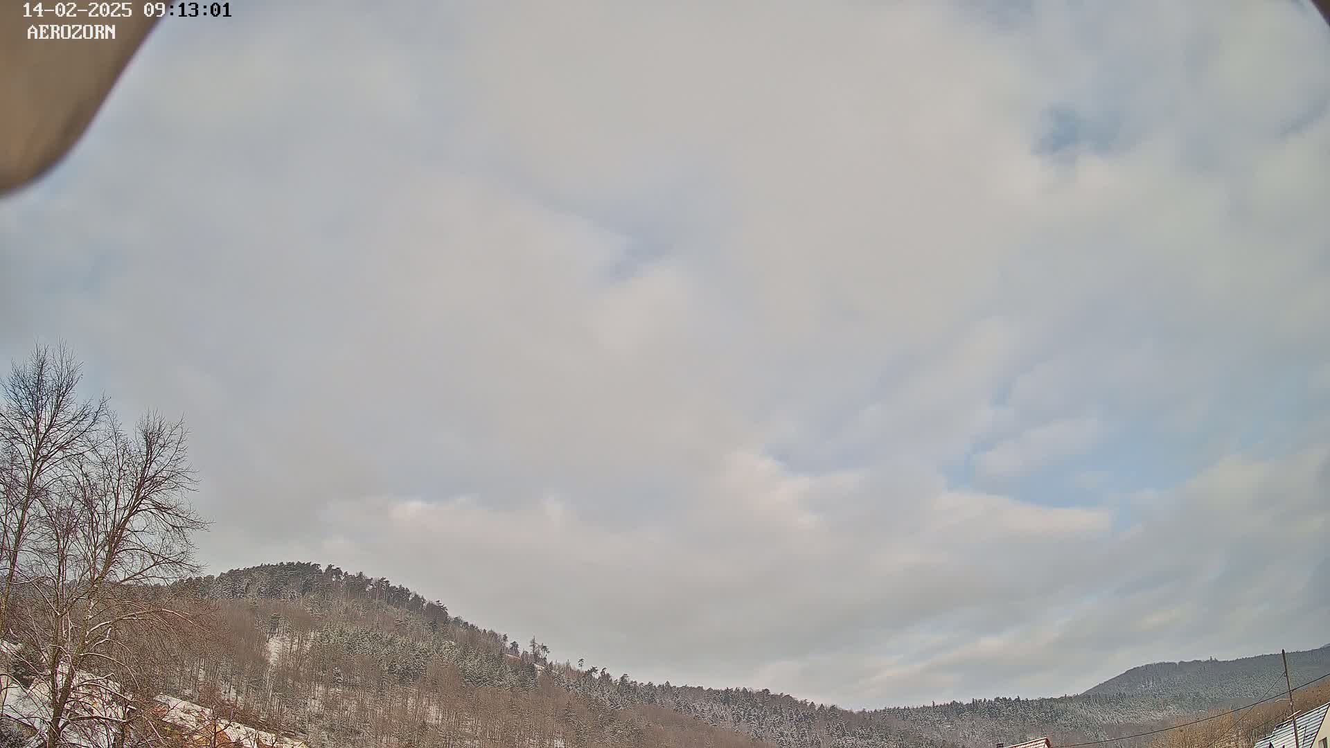 A mostly cloudy sky overlooks snow-covered hills and leafless trees.