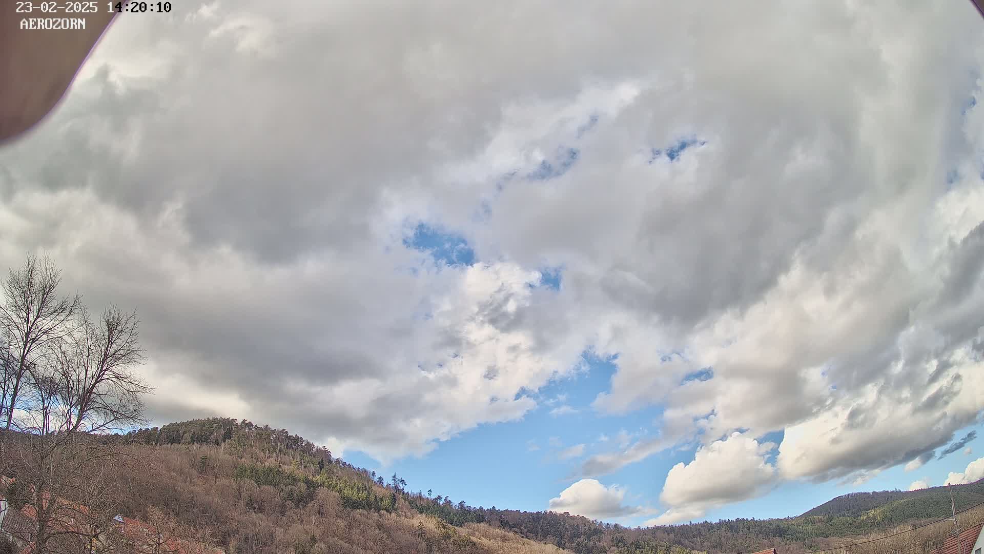 A mostly cloudy sky with patches of blue overhead, above a line of leafless trees and hills.