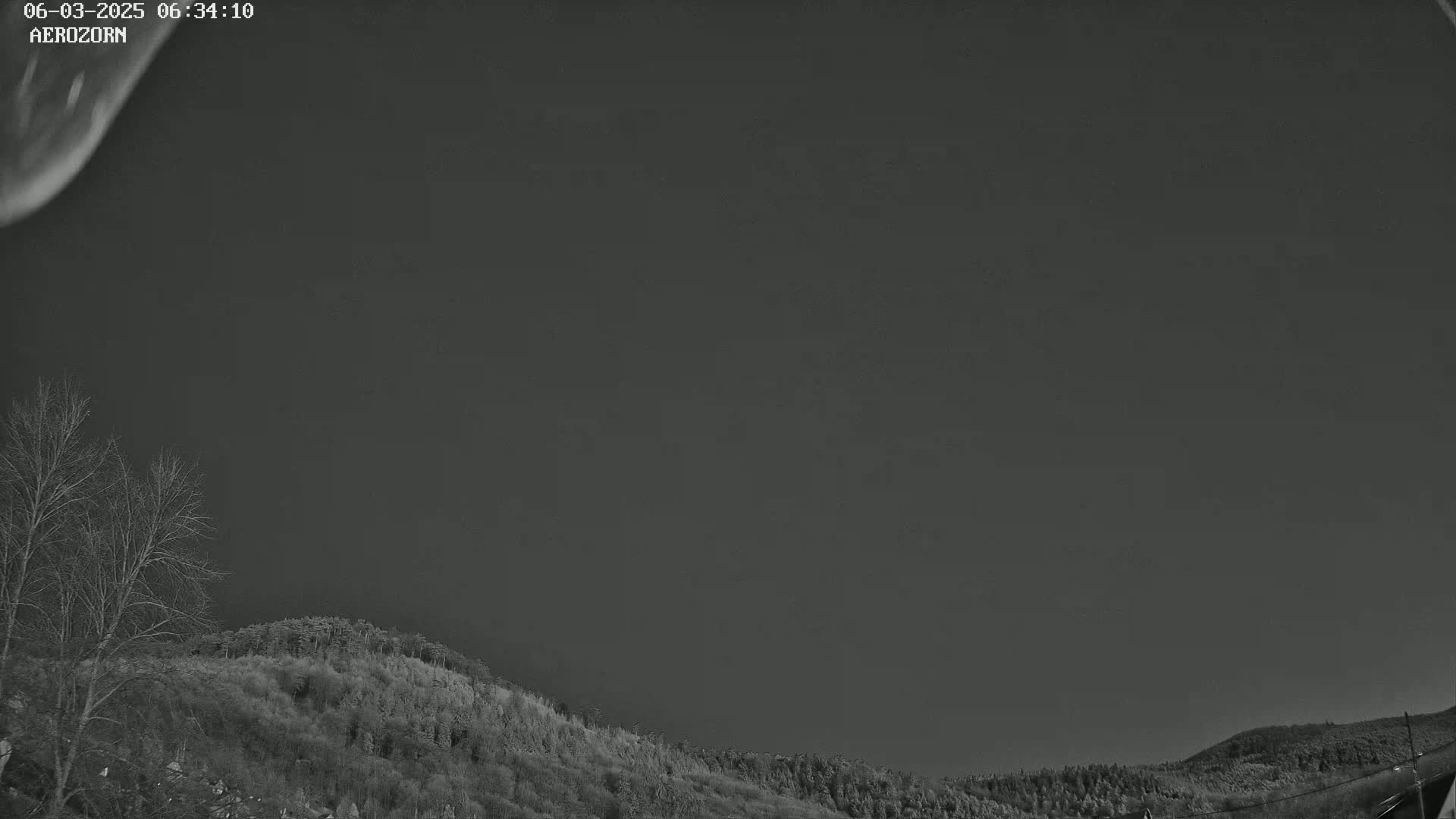 A mostly dark sky over a hillside covered in dark, leafless trees and shrubs.