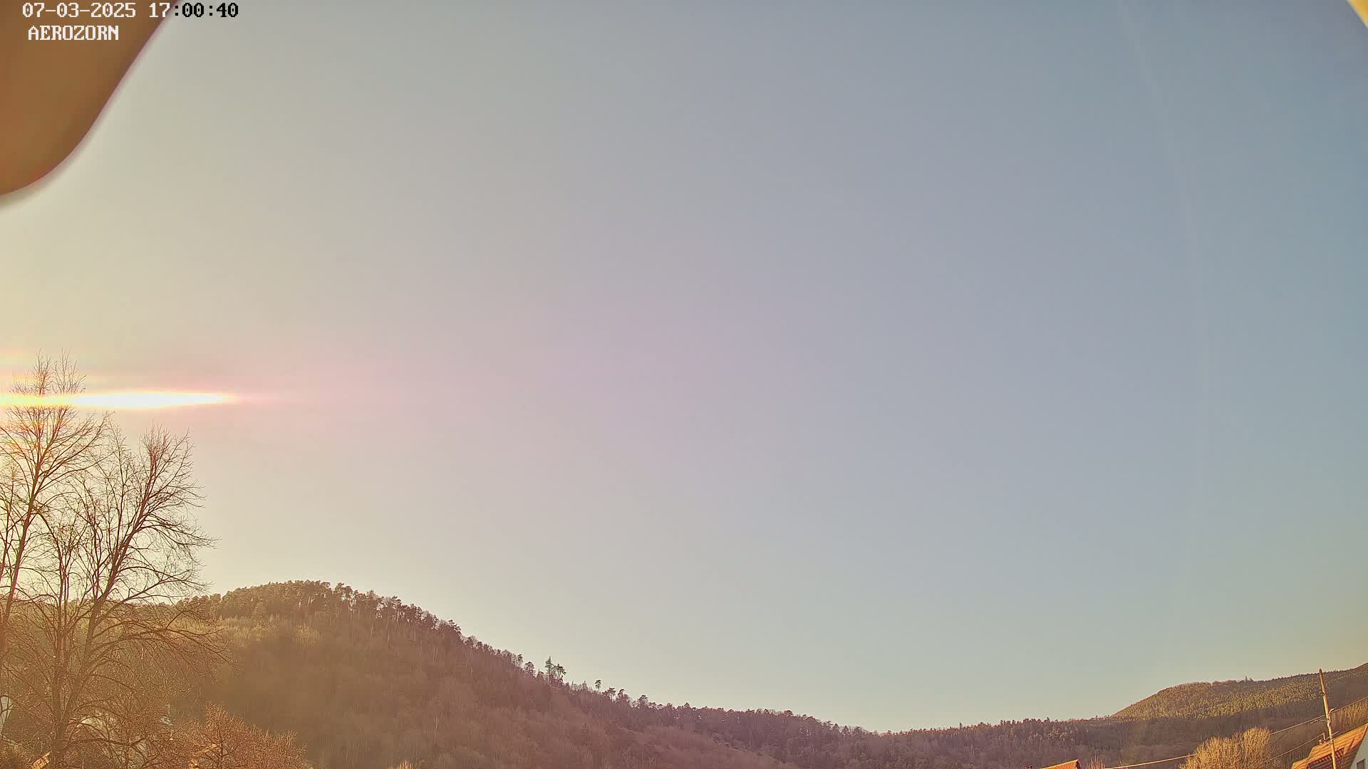 A pale sunset illuminates a mostly clear sky above a line of dark, leafless trees and hills.