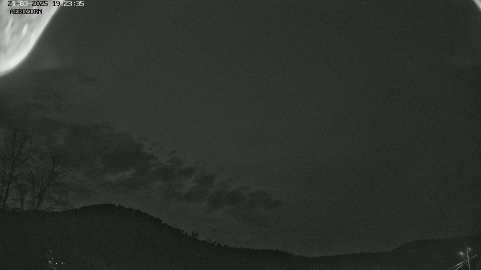 A nighttime view shows a mostly cloudy sky with a faint aurora borealis over a dark silhouetted landscape of trees and hills.