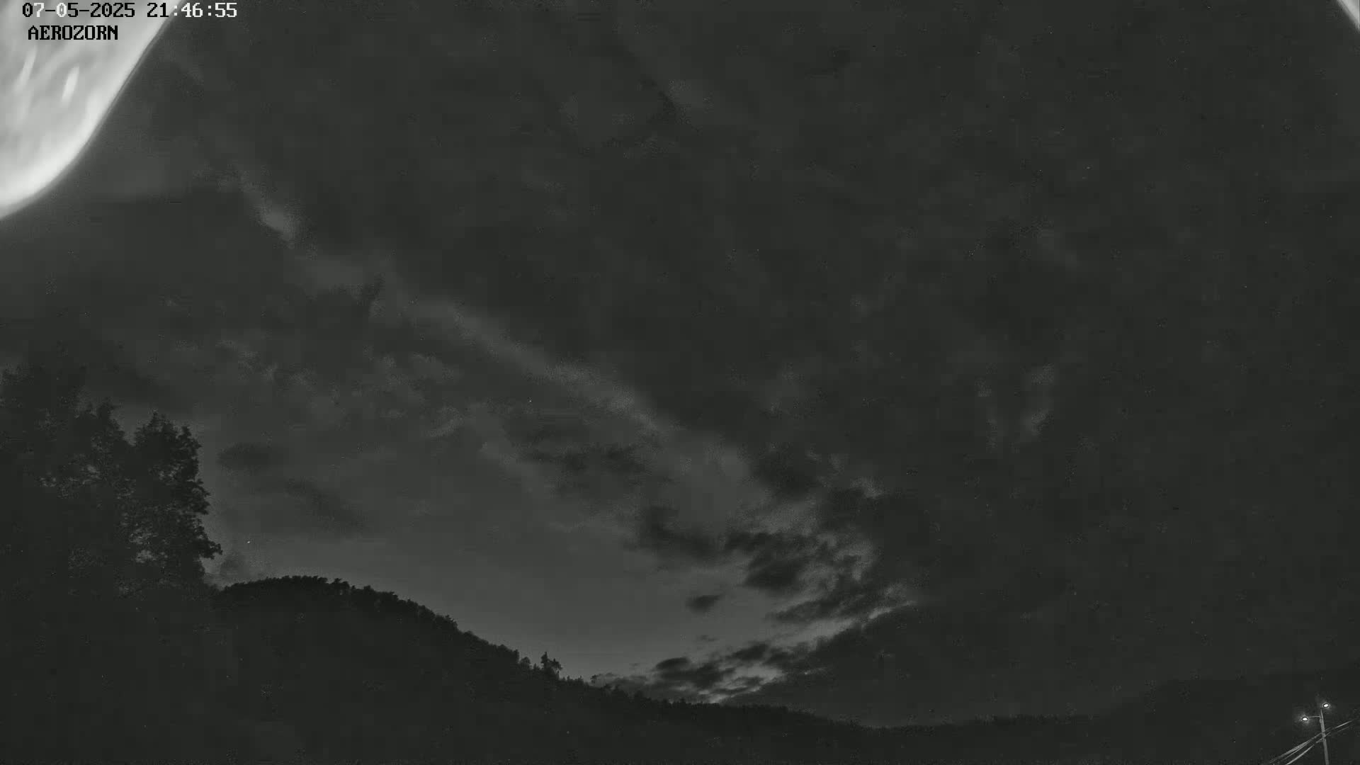 A nighttime, cloudy sky with a faint aurora borealis visible above a dark silhouetted landscape of trees and hills.